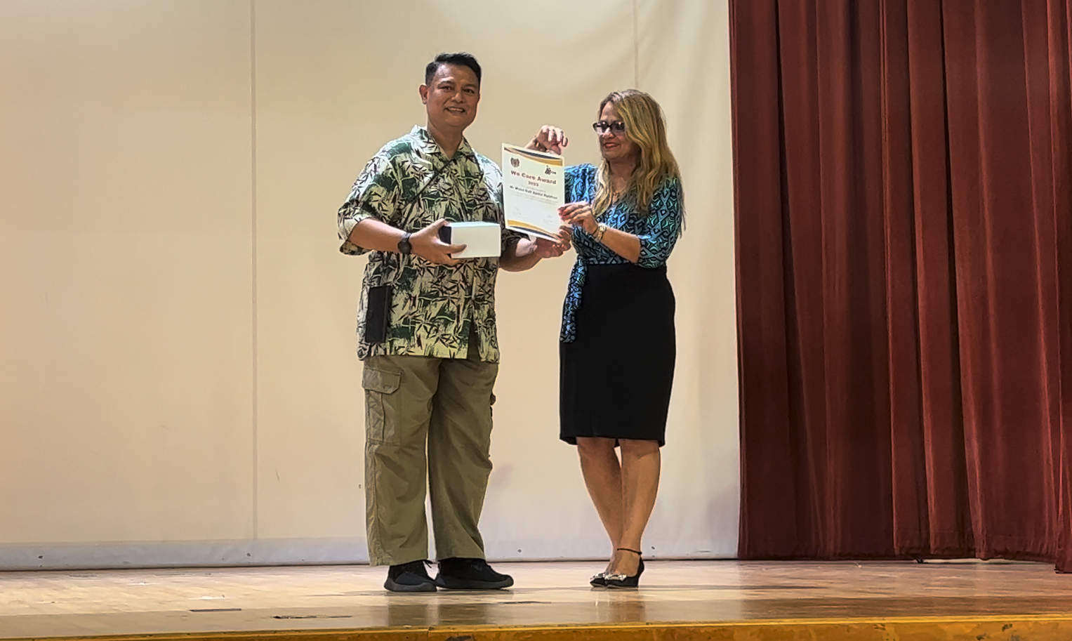 Two people stand on a stage holding a certificate titled We Care Award in front of a red curtain.